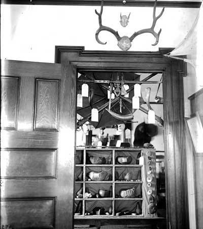An interior photograph of many different kinds of artifacts in a doorway. According to writing on the photograph, these artifacts were used in the Whitman killings in 1847 by some of the Cayuse Tribe. There are Power Horns, Horns, Hatchet Style Pipes, and Rifles. A few of the items have identification tags hanging on the artifacts. Sitting on a table is a box with twelve shelves; inside of each shelf are Stone Bowls and Pestles. On the right side of the box is about a four to five inch space covered with cloth. Hanging on top of the cloth are ten different Fish Weights. In the background are other artifacts, but the photograph is out of focus and it's hard to tell what they are. In front of the doorway on the right side of the photograph is a wooden frame hanging on the wall. 